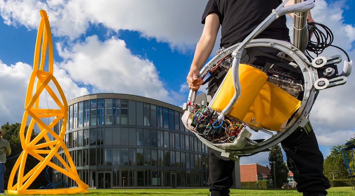 Project number 6. AirTied. A tall yellow inflated truss tower and a person holding the round, metallic AirTied device that fabricated the tower.